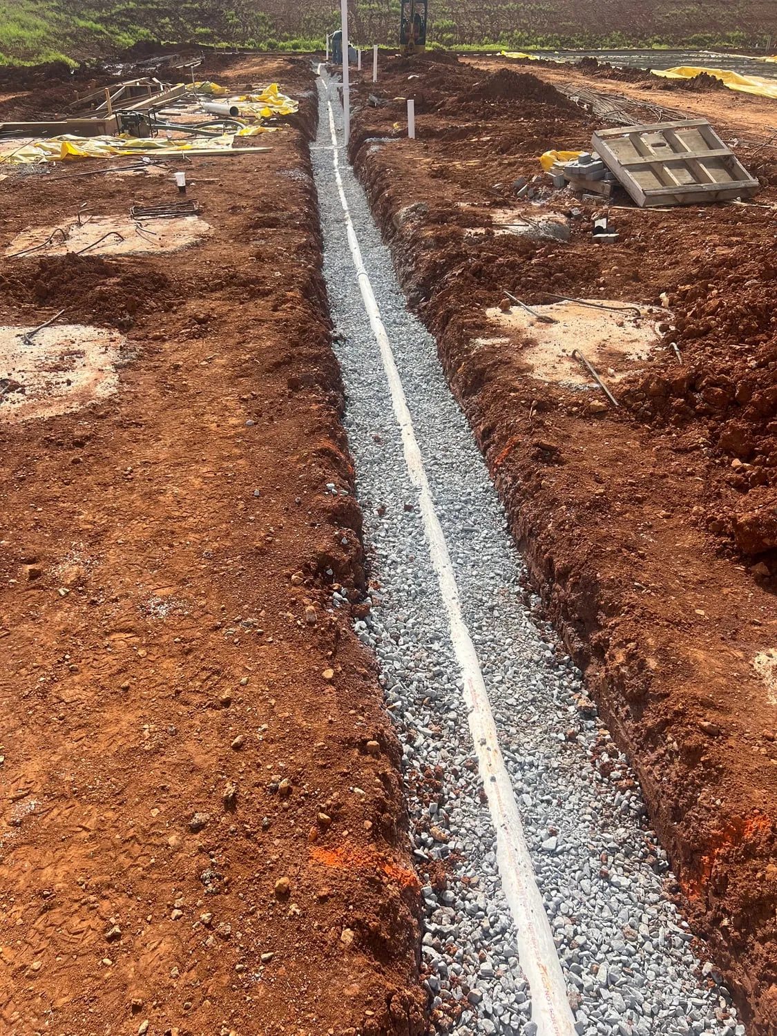 Long trench with new pipe laid on gravel bedding across a red clay lot