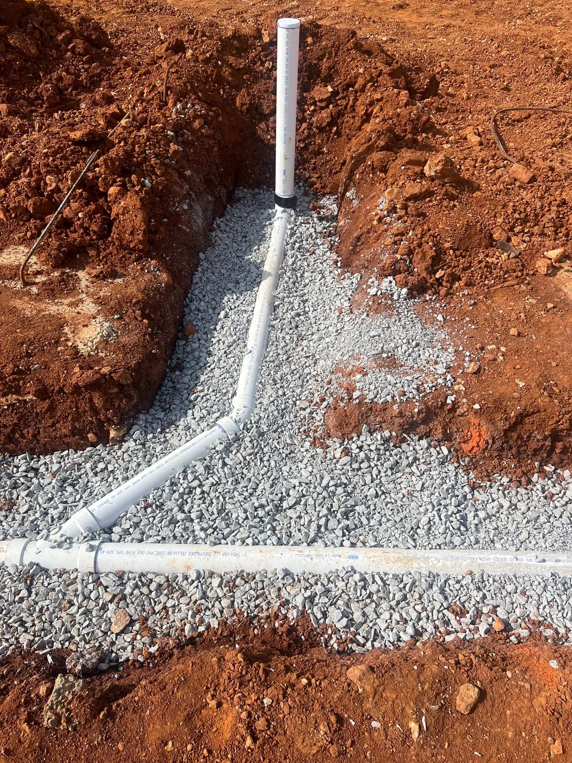 New PVC drain line bedded in gravel in a red clay trench, with a cleanout riser