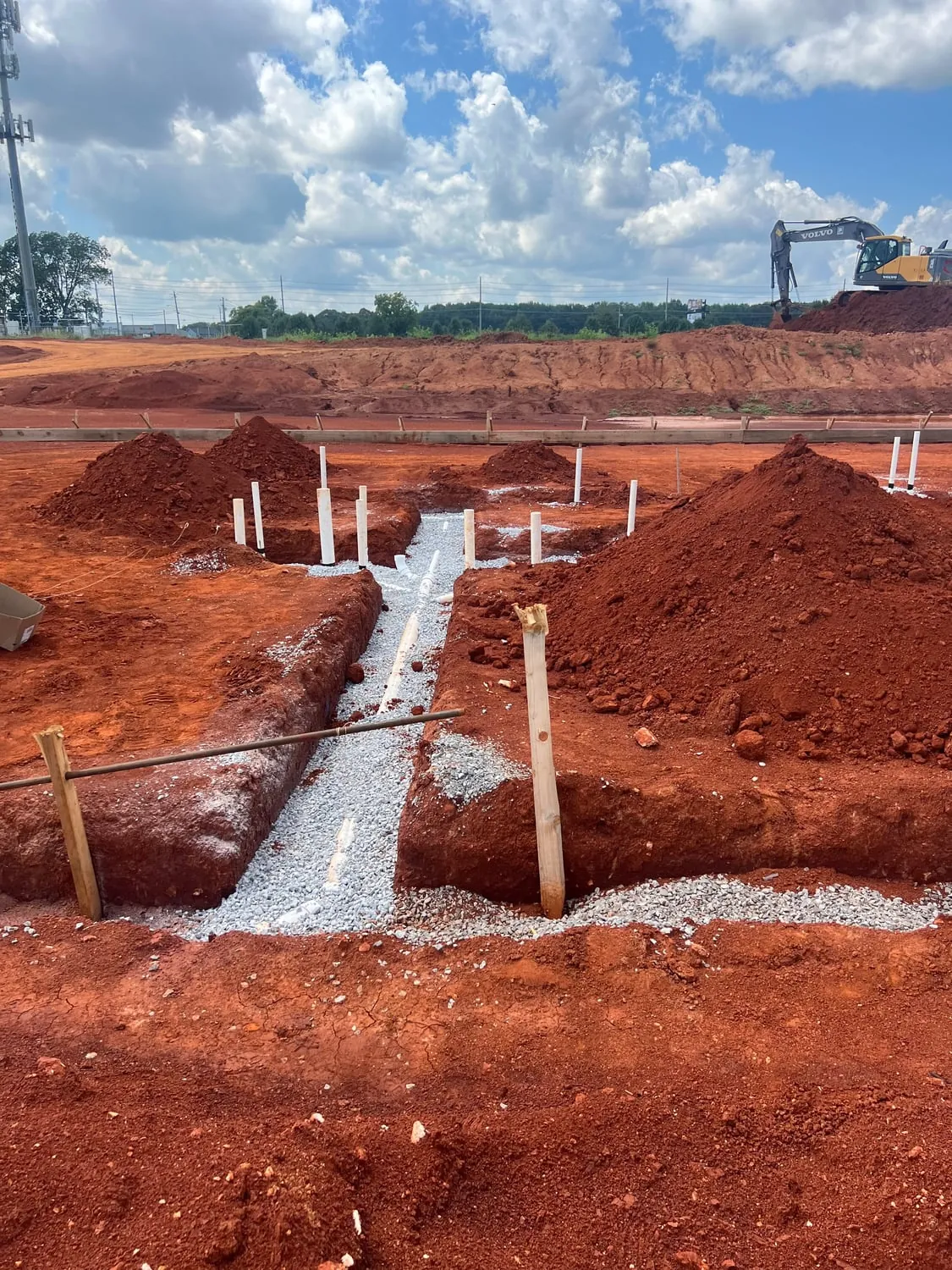Gravel-bedded trench and plumbing stub-ups on a red clay building site with an excavator behind